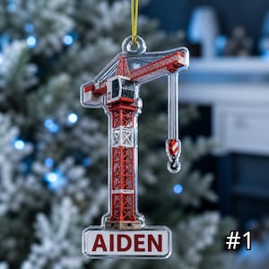 Può includere: Un ornamento sospeso in acrilico trasparente a forma di gru da cantiere. La gru è colorata in rosso e bianco con un gancio nero. Alla base della gru, una placca rettangolare bianca presenta il nome AIDEN stampato in lettere rosse.