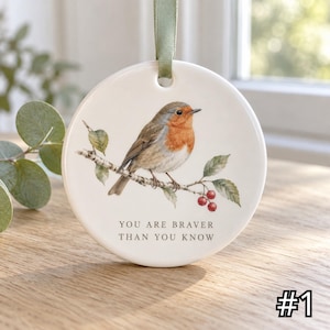 Può includere: Un ornamento rotondo in ceramica bianca appeso a un nastro verde salvia. L'ornamento presenta l'illustrazione di un pettirosso appollaiato su un ramo con foglie verdi e bacche rosse. Sotto l'uccello, il testo recita: YOU ARE BRAVER THAN YOU KNOW.