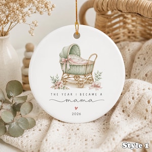 Op de afbeelding: Een rond wit keramieken ornament met een aquarelillustratie van een saliegroene kinderwagen met een roze strik. Onder de kinderwagen staat de tekst THE YEAR I BECAME A mama, gevolgd door een klein roze hartje en het jaartal 2026 in het zwart.