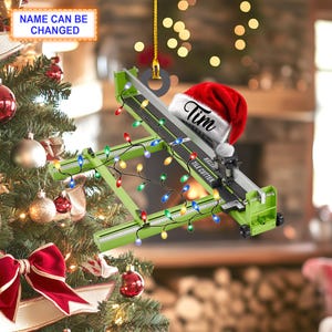 May include: A green tile cutter with a red Santa hat that says "Tim" on it. The tile cutter is hanging from a Christmas tree with a string of colorful lights.
