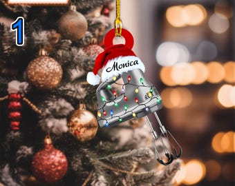 May include: A silver hand mixer Christmas ornament with a red Santa hat and the name "Monica" written on it. The mixer is decorated with colorful Christmas lights.