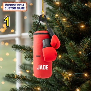 May include: A red boxing-themed Christmas ornament featuring a miniature punching bag and boxing gloves. The ornament has the name "JADE" printed on the bag. The text "CHOOSE PIC & CUSTOM NAME" is at the top. The ornament hangs from a Christmas tree.