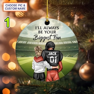 May include: A round, white ornament with a football field background. The ornament features a couple, a woman in a white shirt with the name "Anna" and a man in a black and red football uniform with the name "Jack" and the number "01". The text "I'll Always Be Your Biggest Fan" is printed on the ornament.