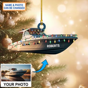 May include: A white and gray boat with colorful lights strung around it. The boat has the name "ROBERTS" on the side. The boat is hanging from a gold string and is a Christmas ornament.