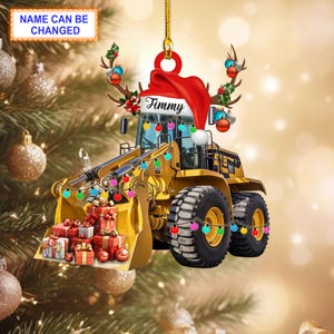 May include: A yellow front-end loader Christmas ornament with a red Santa hat and antlers. The ornament has a string of colorful lights and a bucket filled with wrapped gifts. The text "Timmy" is on the hat, and "Name Can Be Changed" is on a separate label.