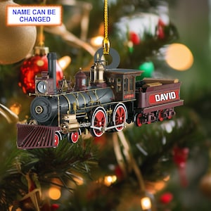 May include: A red, black, and gold toy train ornament with the name "DAVID" on the back. The ornament is hanging from a gold string on a Christmas tree.