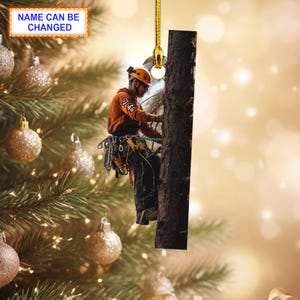 Könnte beinhalten: Ein Weihnachtsbaumschmuck mit einer Person in orangefarbener Kleidung, die an einem Baum klettert. Der Schmuck ist rechteckig und hat eine goldene Schnur, die oben befestigt ist.