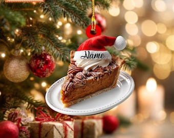 Peut inclure: Ornement de Noël en forme de part de tarte aux noix de pécan, coiffée d'une crème fouettée et d'un bonnet de Père Noël. L'ornement est posé sur une assiette blanche et porte le mot "Name". Arrière-plan avec sapin et cadeaux.