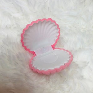 Op de afbeelding: Een roze fluwelen sieradendoosje in de vorm van een schelp, met een wit interieur. De doos is open en het deksel is opengeklapt.