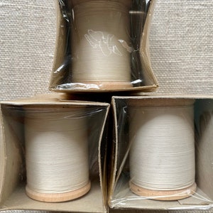 Op de afbeelding: Drie spoelen witte draad verpakt in doorzichtig plastic en bruine kartonnen dozen. De draad is gewikkeld om houten spoelen.