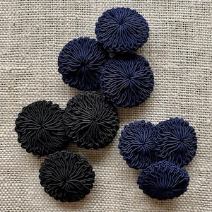May include: A collection of round, circular buttons featuring a dense, radial corded pattern. The set includes several dark navy blue buttons and three solid black buttons, all with a textured, woven surface and a uniform, circular shape.