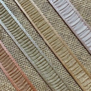 May include: Four different trims of ribbon with a textured pattern. The ribbons are in shades of beige, peach, and light pink. The ribbons are arranged diagonally on a burlap background.
