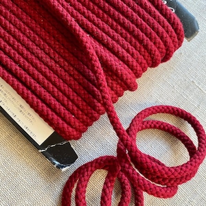 May include: A spool of deep red braided cord wrapped around a black cardboard core. The cord has a thick, textured, woven appearance and is coiled neatly on the spool, with a loose end extending outward and curling into two circular loops.