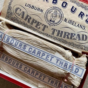 May include: A vintage red box containing bundles of off-white carpet thread. The box lid features the text BARBOUR'S LISBURN N. IRELAND CARPET THREAD and a logo of a hand holding flax. Each bundle is wrapped in a paper label reading BARBOUR'S CARPET THREAD.