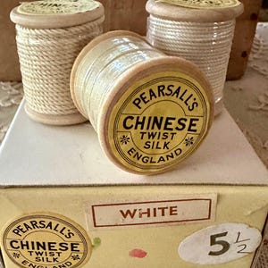 May include: Three wooden spools of white Pearsall's Chinese Twist Silk thread resting on a light yellow cardboard box. The box features a label reading "WHITE" and "5 1/2" alongside a circular logo with the text "PEARSALL'S CHINESE TWIST SILK ENGLAND".