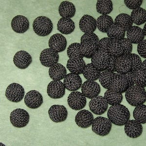 May include: A collection of small, round, black buttons with a textured, crocheted surface. Each button features a dense, woven thread pattern that creates a spherical shape, scattered across the frame in a loose, unorganized arrangement.
