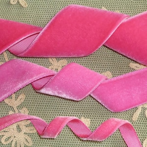 May include: Close-up of three strands of bright pink velvet ribbon. The ribbon is arranged in a wavy pattern, with the edges of the ribbon showing a lighter pink color. The background is a light green fabric with a floral pattern.