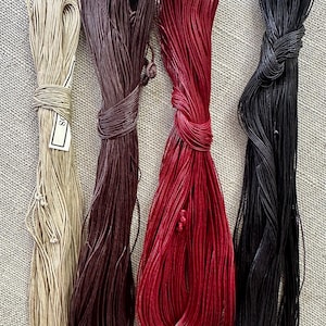 May include: Four bundles of thin, round cord arranged vertically. The colors from left to right are light beige, dark brown, bright red, and black. Each bundle is tied with a small knot near the top, and the beige bundle features a small white label.