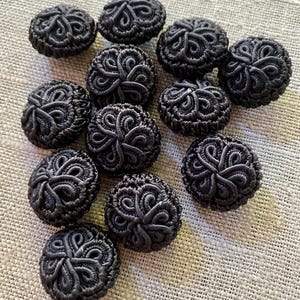May include: A collection of eleven round black buttons, each featuring a raised, intricate corded pattern that forms a symmetrical, swirling design across the surface. The buttons have a textured, fabric-covered appearance with a consistent dark finish.