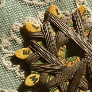 May include: A vintage star-shaped cardboard spool wrapped in dark brown thread. The yellow cardboard features black lettering spelling SEWELL and the text BRITISH MADE. The spool has multiple points and a central circular design with green accents.