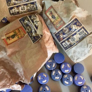 Può includere: Diversi pacchetti in carta marrone etichettati Fil En Capsule Au Conscrit con adesivi rettangolari blu e bianchi, insieme a numerose piccole bobine rotonde in plastica blu contenenti filo bianco, sparse sulla superficie.
