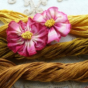 May include: Two fabric flowers with pink and white striped petals and bright yellow knotted centers, resting on top of multiple skeins of golden-yellow and mustard-colored embroidery thread, featuring a textured, layered appearance.