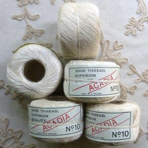 Peut inclure: Cinq bobines de fil à chaussures blanc cassé disposées en groupe. Chaque bobine porte une étiquette en papier avec le texte "SHOE THREAD, SUPERIOR QUALITY", "ACADIA" et "No 10", ainsi que la mention "MADE IN N. IRELAND" imprimée en noir et rouge.