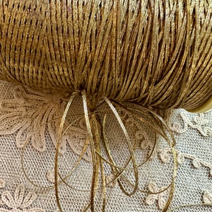 May include: A spool of gold metallic cord, with a few strands of the cord unraveled and lying on a beige lace fabric.