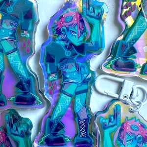 May include: A holographic charm featuring a cartoon character with pink hair, a black shirt, and blue jeans. The character is holding a gun and has a skull and crossbones on their shirt. The charm is attached to a white metal keychain.