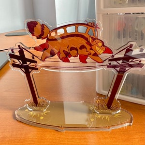 May include: A clear acrylic stand featuring a cartoon cat with a bus on its back, walking on power lines. The cat is brown and orange with black stripes. The bus is yellow with brown stripes. The stand has a brown base with green grass and yellow flowers.