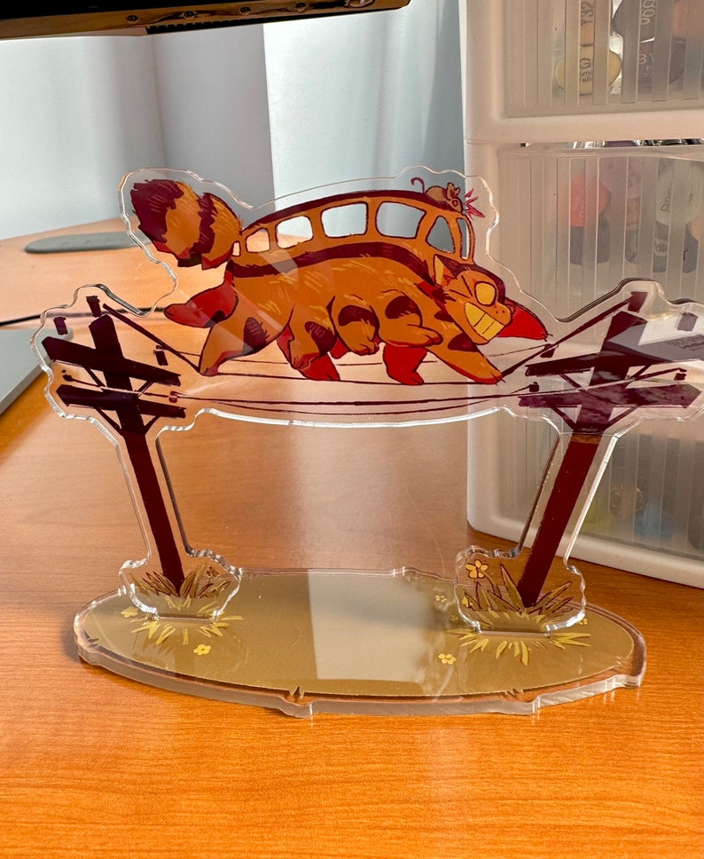 May include: A clear acrylic stand featuring a cartoon cat with a bus on its back, walking on power lines. The cat is brown and orange with black stripes. The bus is yellow with brown stripes. The stand has a brown base with green grass and yellow flowers.