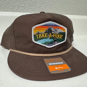 Pode incluir: Boné de beisebol castanho com aba plana e detalhe de corda. O boné apresenta um patch bordado com o texto "TAKE A HIKE" sobre uma cena de montanha e lago. Uma etiqueta com a marca "RICHARDSON" está anexada.