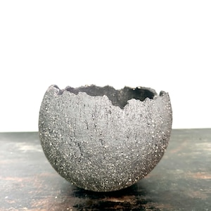 Könnte beinhalten: Eine dunkelgraue, strukturierte Keramikschale mit einem rauen, unebenen Rand. Die Schale hat eine kugelförmige Form und eine gesprenkelte Oberfläche, die ihr ein rustikales Aussehen verleiht. Die Schale steht auf einer dunklen Holzoberfläche.