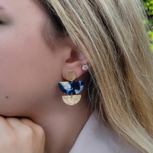 May include: A pair of gold and blue statement earrings with a geometric design. The earrings feature a large, semi-circular blue and white patterned piece with a smaller gold disc above it. The earrings are worn by a person with blonde hair.