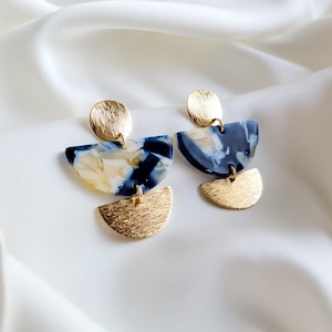 May include: A pair of gold and blue statement earrings with a geometric design. The earrings feature a large, semi-circular blue and white patterned piece, a smaller gold semi-circle, and a round gold piece at the top.
