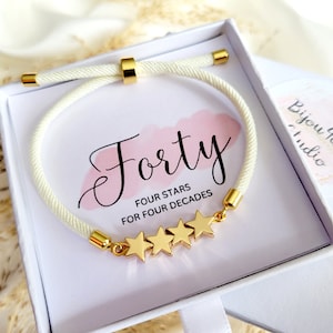 May include: White cord bracelet with gold stars and the text "Forty Four Stars For Four Decades" on a white box with the text "Bijou Hall Studio".