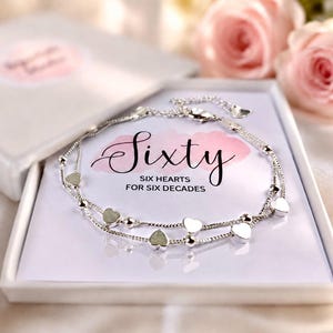 May include: A silver-toned double-strand bracelet featuring six small heart charms, presented on a white card with the text "Sixty" and "SIX HEARTS FOR SIX DECADES" printed in black, enclosed in a white gift box with a matching lid.
