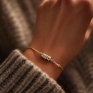 May include: A gold-toned bracelet featuring a thin, delicate chain and a central focal point consisting of several small, textured, cylindrical gold-toned beads clustered together on the chain, creating a subtle and refined metallic accessory.