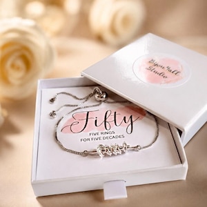 May include: A silver-toned adjustable bracelet featuring five small rings on a central bar, presented in a white gift box. The box insert displays the text Fifty, FIVE RINGS FOR FIVE DECADES, and the box lid features a Byou'tall Studio sticker.