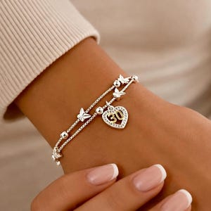 May include: A double-strand silver-toned bracelet featuring small metallic butterfly charms and spherical beads. A heart-shaped pendant hangs from the bracelet, containing the number 30 in gold-toned metal surrounded by clear crystals.