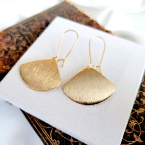 May include: A pair of gold-toned earrings with a fan-shaped design. The earrings have a brushed finish and are suspended from thin, gold-toned wires.