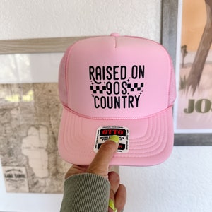 K&ouml;nnte beinhalten: Rosafarbener Trucker-Hut mit schwarzem Netzr&uuml;cken und schwarzem Text, der "RAISED ON 90S COUNTRY" mit einem karierten Muster lautet.