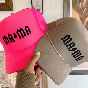K&ouml;nnte beinhalten: Zwei Trucker-Caps mit Netzr&uuml;cken. Eine M&uuml;tze ist pink mit schwarzem Text, der "mama" mit einem Blitz sagt. Die andere M&uuml;tze ist braun mit schwarzem Text, der "mama" mit einem Blitz sagt.