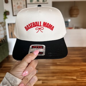May include: White baseball cap with black brim. The front of the cap has red embroidery that reads "Baseball Mama" with a red bow.