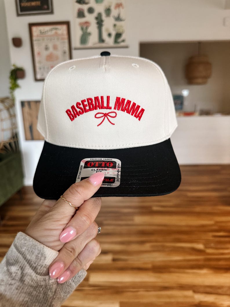 May include: White baseball cap with black brim. The front of the cap has red embroidery that reads "Baseball Mama" with a red bow.