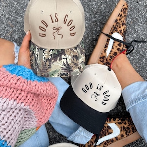 Op de afbeelding: Twee baseballpetten met de tekst "God is Good" geborduurd op de voorkant. De ene pet is beige met een camouflagekleurige rand en de andere is zwart met een witte rand.