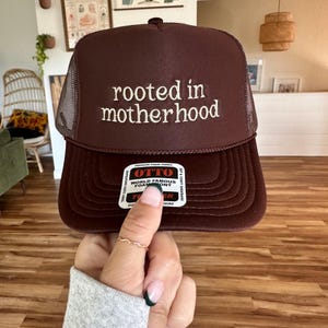 May include: A brown trucker hat with a mesh back. The hat has white embroidery that reads "rooted in motherhood".