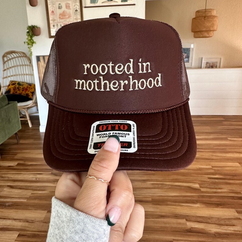 May include: A brown trucker hat with a mesh back. The hat has white embroidery that reads "rooted in motherhood".