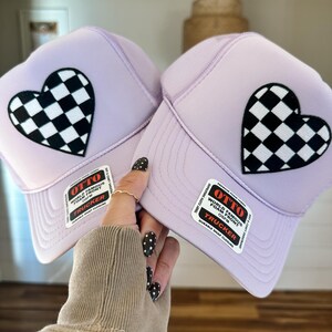 May include: Two lavender trucker hats with a black and white checkered heart patch. Each hat has a white tag with the text "OTTO WORLD FAMOUS FOAM FRONT OSFM TRUCKER DURABLE SNAP CLOSURE".