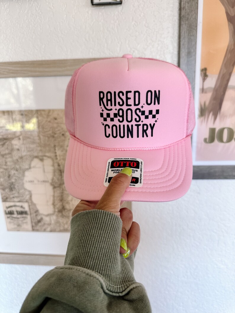 K&ouml;nnte beinhalten: Rosafarbener Trucker-Hut mit schwarzem Netzr&uuml;cken und schwarzem Text, der "RAISED ON 90S COUNTRY" mit einem karierten Muster lautet.